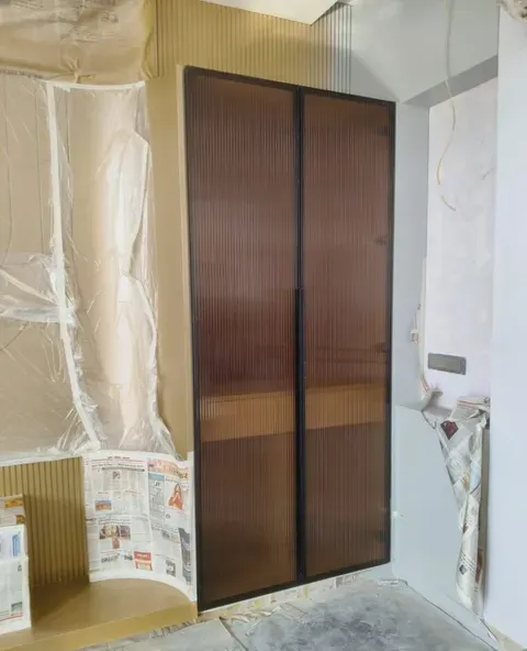 Tall two-door unit with black profile frames and bronze-tinted fluted glass, photographed during installation with the surrounding room still masked